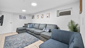 A modern living room with a grey sectional sofa, a blue armchair, a patterned rug, and a potted plant. - Huge Home | Sleeps 10+ | Minutes from NYC (Bayonne)