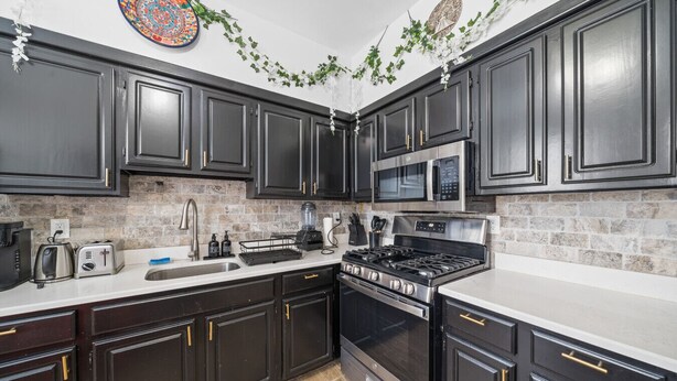 A kitchen with dark cabinets, a stainless steel stove, a microwave, and a brick backsplash. - Huge Home | Sleeps 10+ | Minutes from NYC (Bayonne)