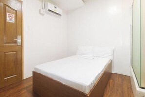 Una habitación pequeña con cama individual, puerta de madera, pared blanca y unidad de aire acondicionado blanca.