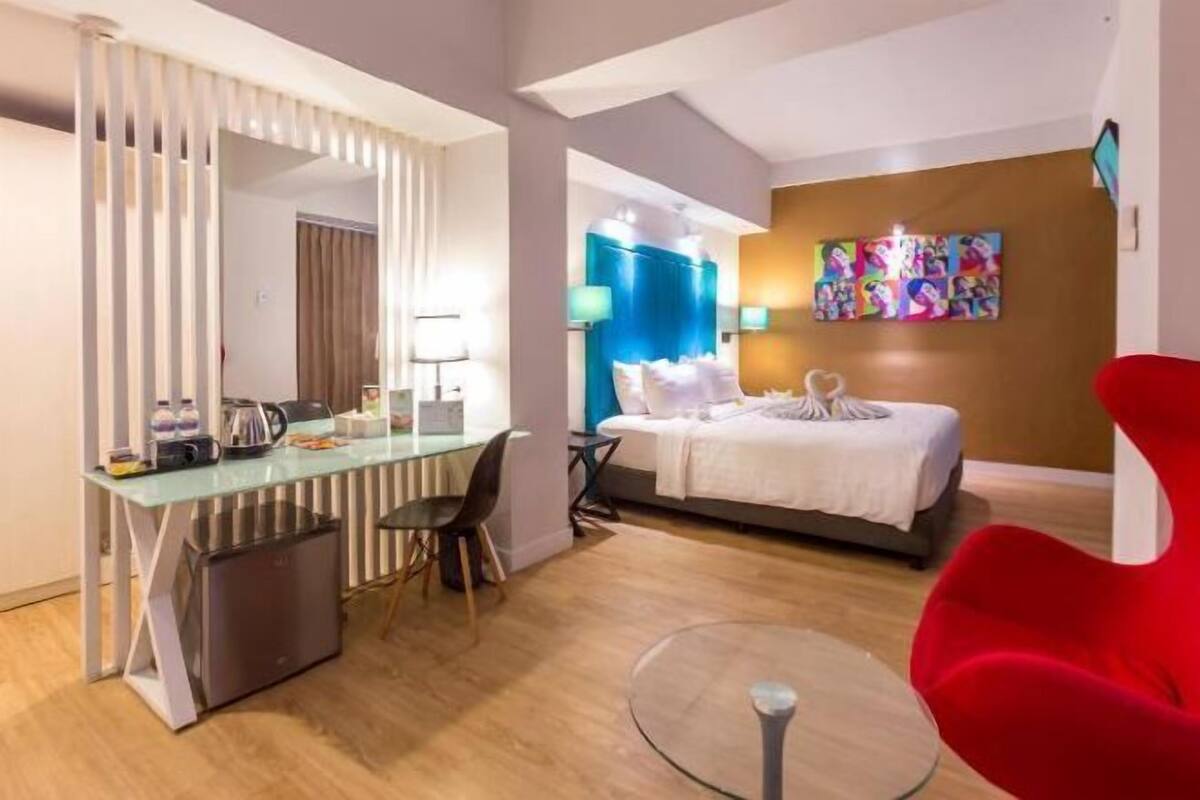 Una habitación de hotel moderna con un escritorio de vidrio, una cama con ropa blanca, una silla roja y una obra de arte colorida en la pared.