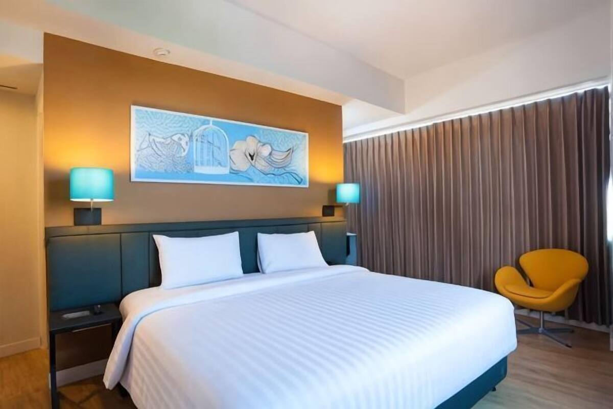 Habitación de hotel con una cama grande, dos lámparas de noche, un cuadro en la pared, una silla amarilla y una ventana con cortinas.