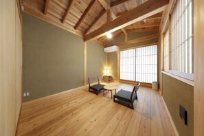 Una habitación amplia con suelo de madera, techo bajo y un gran ventanal con persianas verticales.