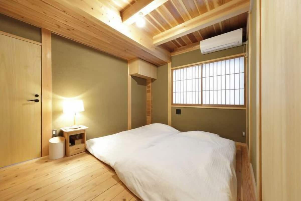 A room with a bed, a nightstand, a lamp, a window with bars, and a wooden ceiling.