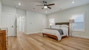 Un dormitorio con piso de madera, una cama con ropa blanca, un ventilador de techo y una lámpara de noche.