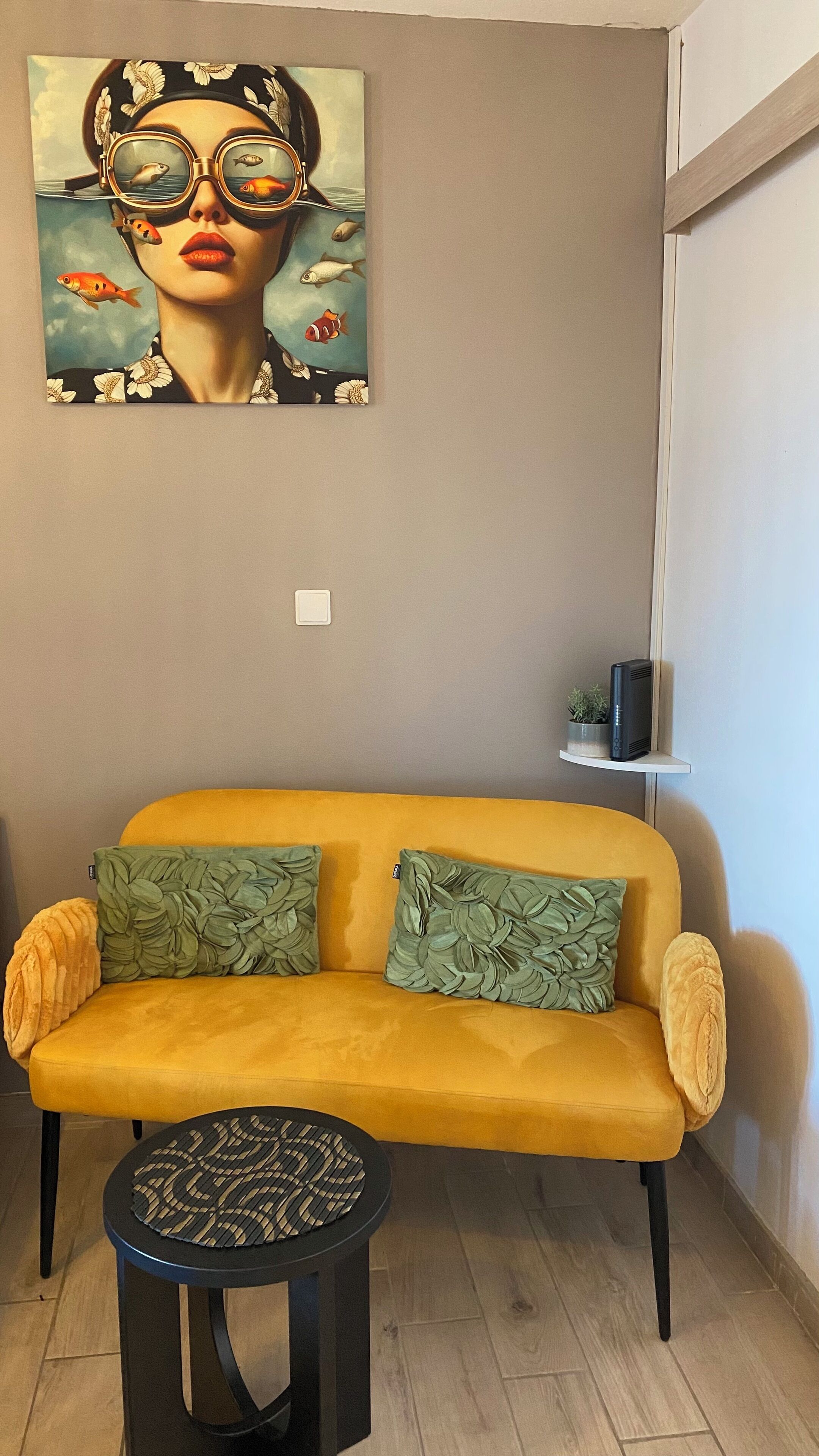 Un sofá amarillo con cojines de estampado de hojas verdes, una pequeña mesa redonda con tapa de mimbre y un cuadro de una mujer con peces en la pared.