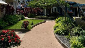 A pathway leading through a courtyard with tropical plants and palm trees, surrounded by colorful buildings with balconies and a clear blue sky. - Les Alizés Joli Studio sea view beach walk Anse Mitan Trois ILETS (Trois Ilets)