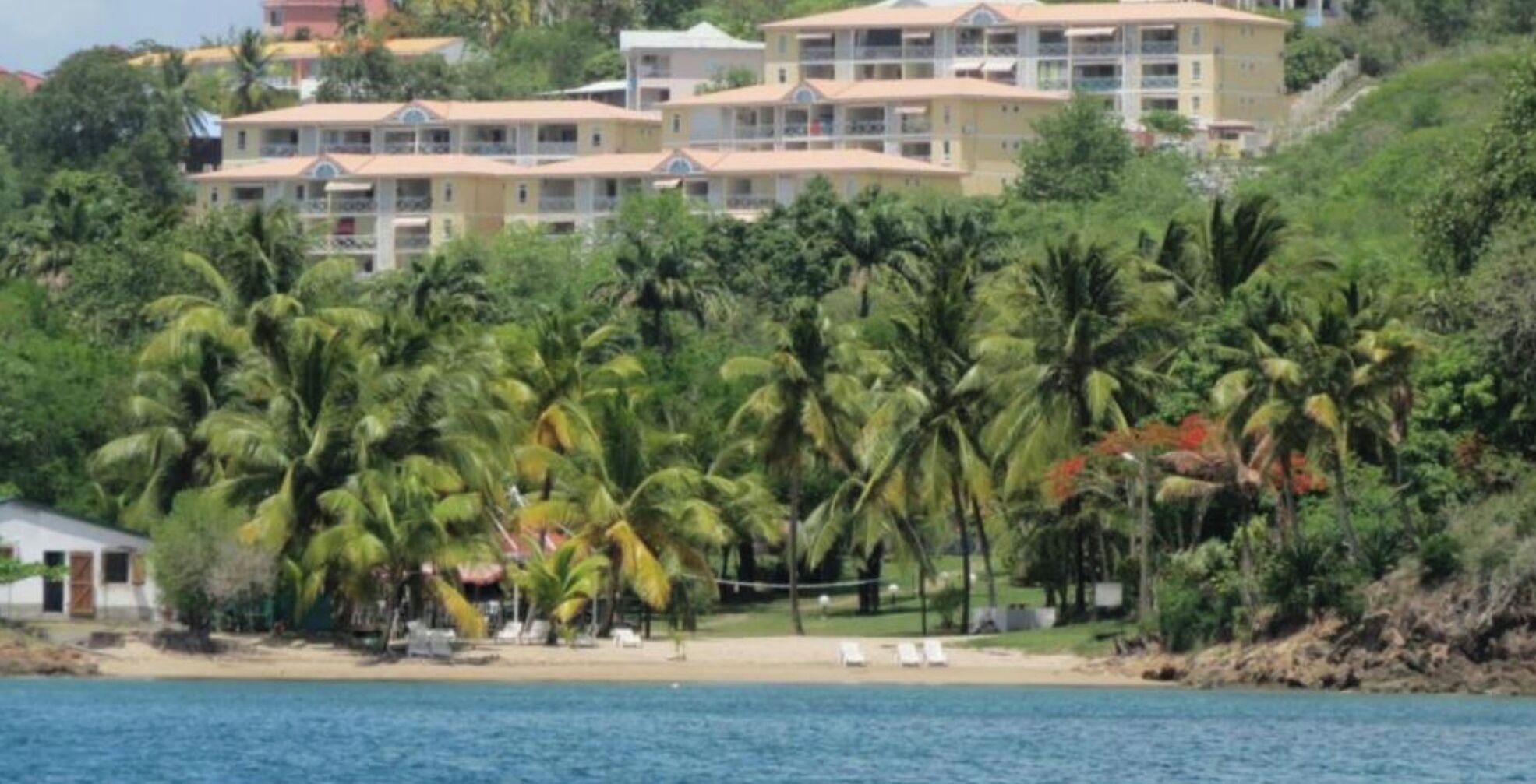Un hotel frente a la playa con varias edificaciones, palmeras y una playa de arena.