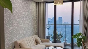 A modern living room with a sofa, a coffee table, and a view of a cityscape through large windows. - Flora Urban - Premium Apartment Vinhomes Skylake Near Keangnam (Hanoi)