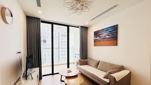 A modern living room with a sofa, a small table, a flat-screen TV, and a large window with curtains. - Flora Urban - Premium Apartment Vinhomes Skylake Near Keangnam (Hanoi)