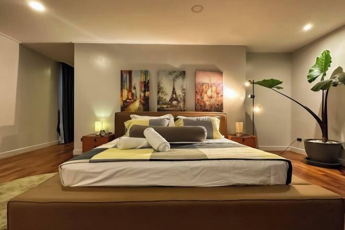 A hotel room with a large bed, a nightstand, a lamp, a potted plant, and three paintings on the wall.