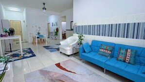 Una sala moderna con un sofá azul, un sillón blanco y una mesa de centro. Se observa un comedor con mesa y sillas, y una cocina al fondo.