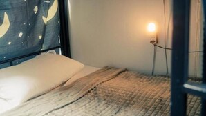 Una cama con un almohadón blanco y una colcha con textura, un enchufe con una bombilla, y una cortina con estampado de luna creciente.