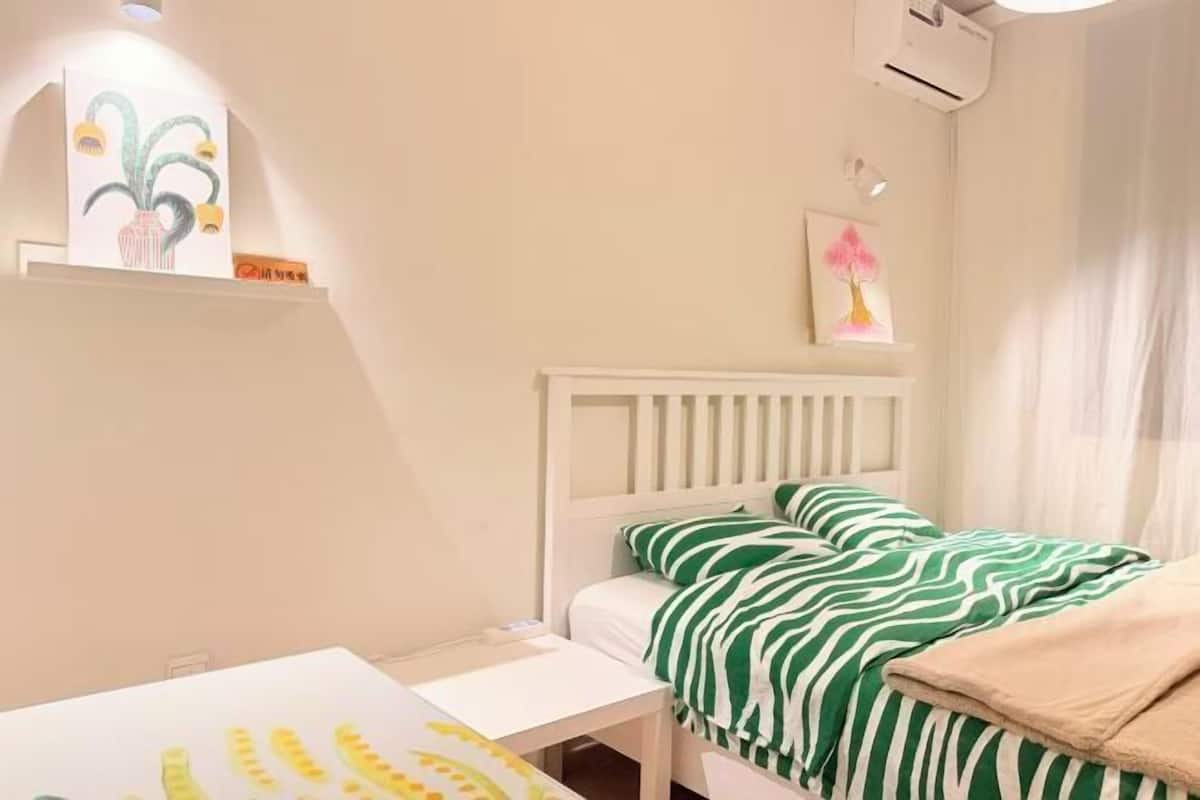 A bedroom with a white bed, green and white striped bedding, a white bedside table, and a wall-mounted shelf with artwork.