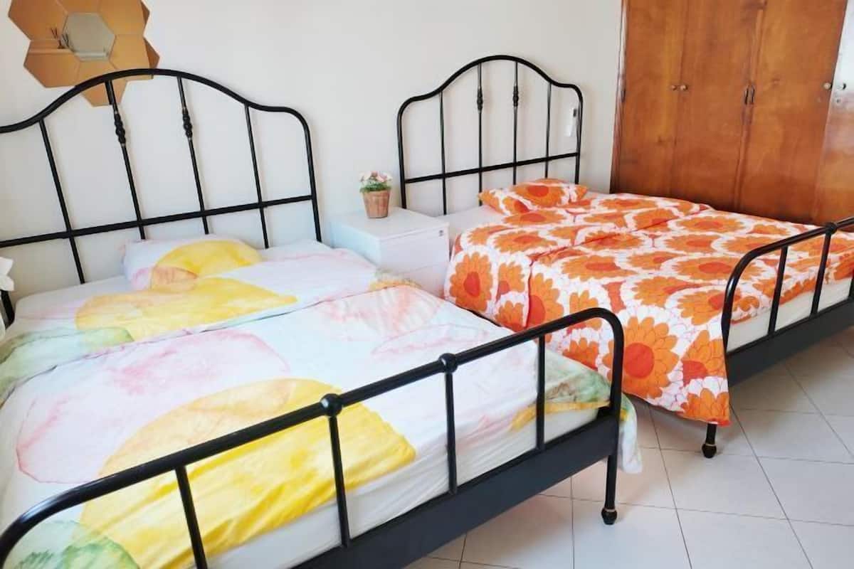 A room with two beds, each with a distinct floral bedspread, a small bedside table with a plant, and a wooden wardrobe.