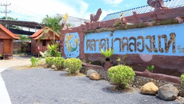 A sign with Thai script, surrounded by rocks and plants, with a traditional roofed building in the background. - Talad Nam Klong Hae Resort (Hat Yai)