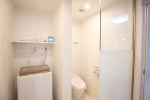 A compact bathroom with a washing machine, toilet, and a small shelf. - Lala Court Seaside Yagaji (Nago)