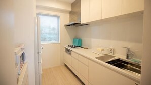 A compact kitchen with white cabinets, a stove, a sink, and a window with blinds. - Lala Court Seaside Yagaji (Nago)