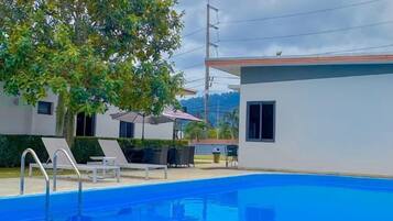 A swimming pool with lounge chairs and umbrellas, surrounded by a building with a flat roof and a tree.