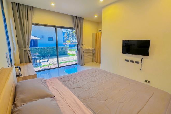 A hotel room with a bed, a TV, a sliding glass door to a balcony, and a small table with a kettle.