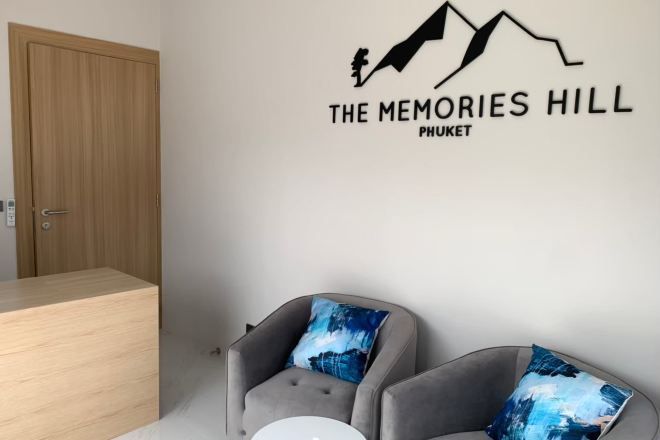 A room with a wooden door, two grey armchairs with blue cushions, and a wall decal that reads 'THE MEMORIES HILL PHUKET'.