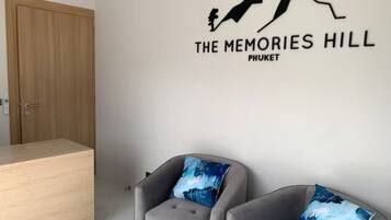 A room with a wooden door, two grey armchairs with blue cushions, and a wall decal that reads 'THE MEMORIES HILL PHUKET'.