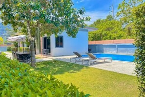 Ett modernt hus med en swimmingpool, en uteplats med stolar och en liten trädgårdsdel.