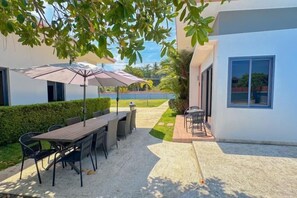 Un patio exterior con una mesa y sillas para comer, rodeado por un seto y un edificio blanco.