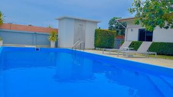 A swimming pool with a clear blue water, surrounded by a white building, a small structure, and a tree.