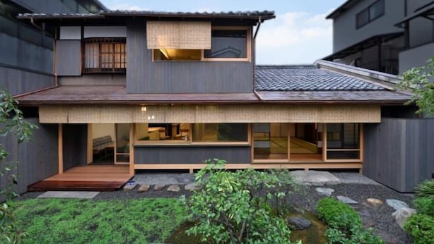 Une maison moderne à deux étages, dotée d’une terrasse en bois, de grandes fenêtres et d’un petit jardin avec un bassin.