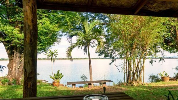 Um deck de madeira com um banco e um objeto de vidro, com vista para um lago, árvores e um céu claro.