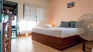Quarto com cama, um ventilador ao lado da cama, janela com cortinas, quadro na parede e porta que dá para outro cômodo.