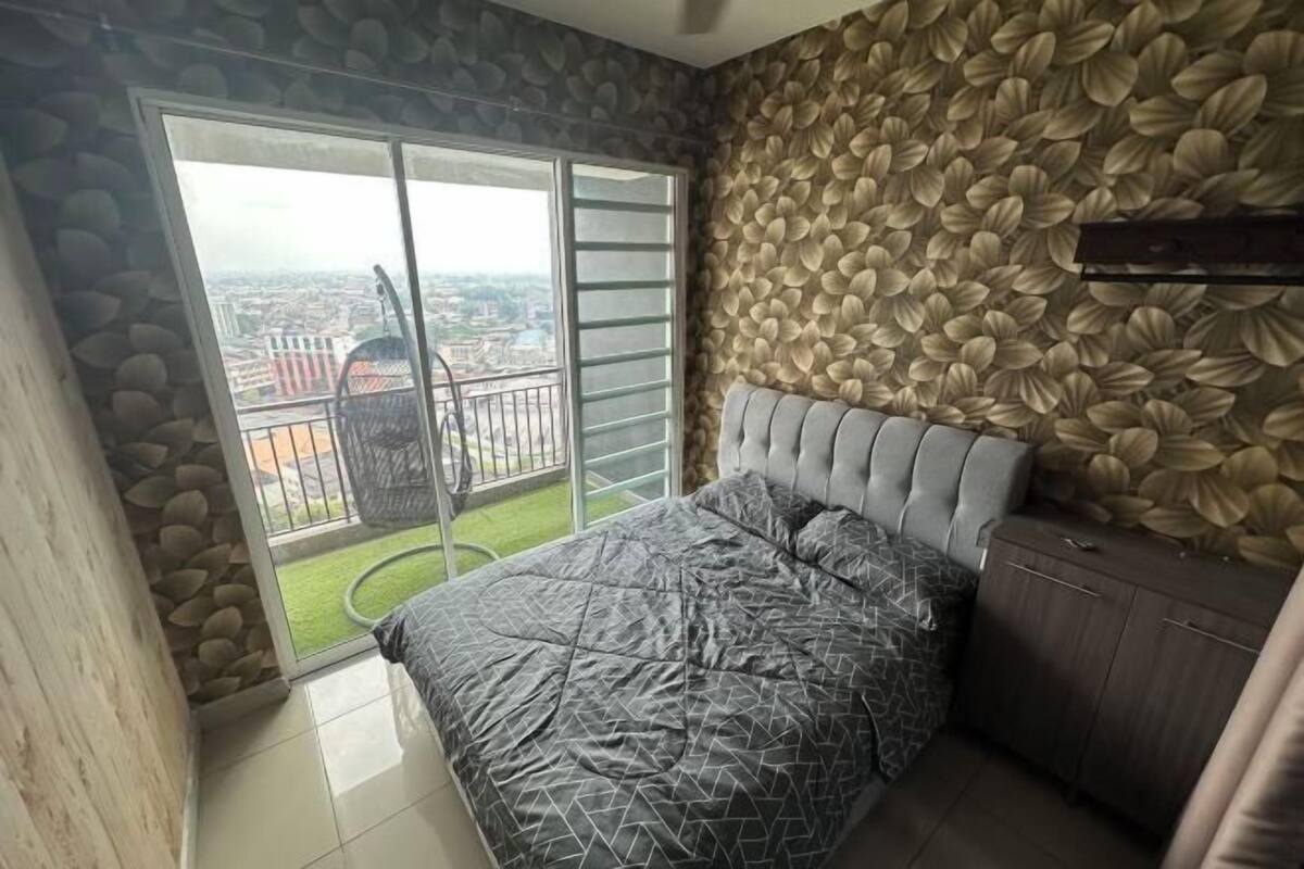 A bedroom with a large sliding glass door, a bed with a patterned bedspread, a grey upholstered headboard, and a wall with a leaf pattern.