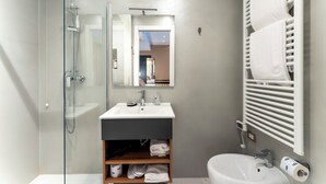 Un bany modern amb dutxa de vidre, un lavabo blanc i un mirall a sobre. Hi ha un suport de tovalloles amb tovalloles i un dispensador de paper higiènic a la paret.