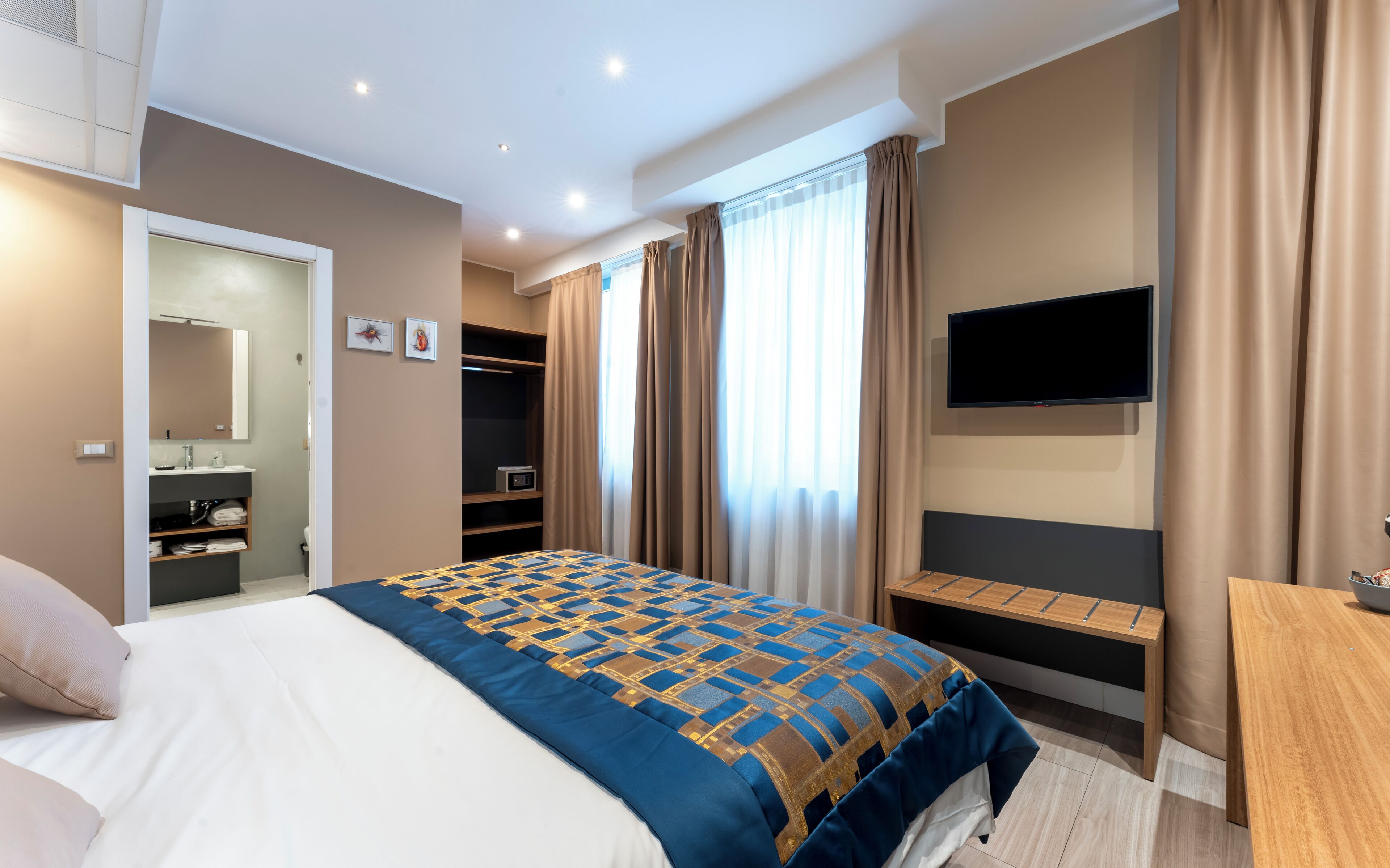 A modern hotel room with a large bed, a TV mounted on the wall, a walk-in closet, and a bathroom with a mirror and sink.