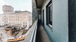 Un balcó estret amb vista a un paisatge urbà, incloent un edifici gran amb moltes finestres i un tramvia en primer pla.