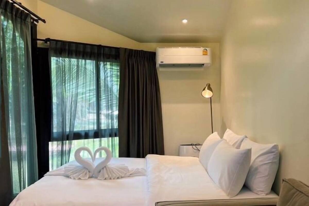 A hotel room with a bed, a wall-mounted air conditioner, a bedside lamp, and a window with curtains.