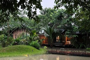 A traditional wooden building with a pond in front, surrounded by lush greenery. - Breathe - Rice Valley (San Sai)