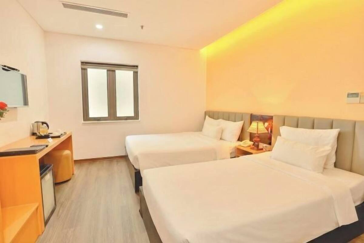 A hotel room with two beds, a desk, a chair, a small table, a lamp, and a window.