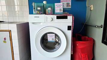 A Samsung washing machine in a laundry room with a blue wall, a red trash bag, and a door with a handle.