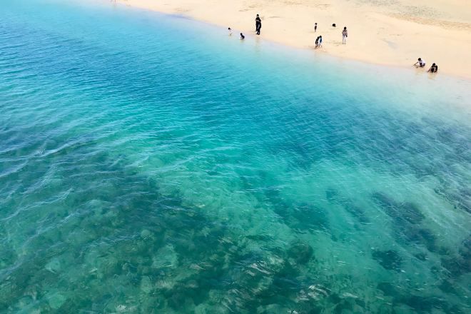 A beach with clear turquoise water and people swimming and walking on the shore.
