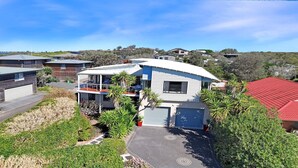 A modern two-story house with a balcony, surrounded by lush greenery and a clear blue sky. - The Break – space, comfort and seaside fun (Culburra Beach)