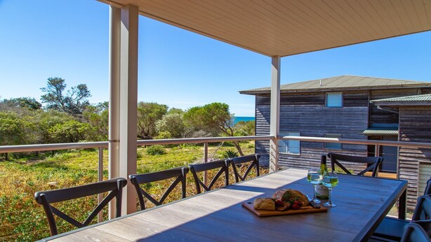 A wooden deck with a table set for dining, overlooking a natural landscape and a wooden building. - The Break – space, comfort and seaside fun (Culburra Beach)