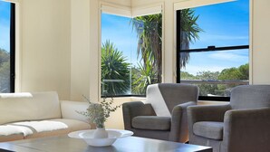 A modern living room with a beige sofa, two grey armchairs, and a coffee table with a white vase. Large windows offer views of palm trees and a clear blue sky. - The Break – space, comfort and seaside fun (Culburra Beach)