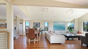 A spacious living area with a wooden dining table, white sofa, and a large painting of a wave. - The Break – space, comfort and seaside fun (Culburra Beach)