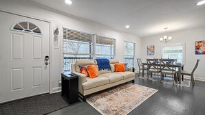 A living room with a beige sofa, a dining table, and chairs. There is a door with a window, and a chandelier on the ceiling. - Walk to AT&T Stadium! | Dream City Retreat | 3BR/2BA w/Firepit & Modern Comforts (Arlington)