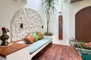 Un espace de détente avec un mur en pierre, un banc en bois agrémenté de coussins, une plante et une piscine.