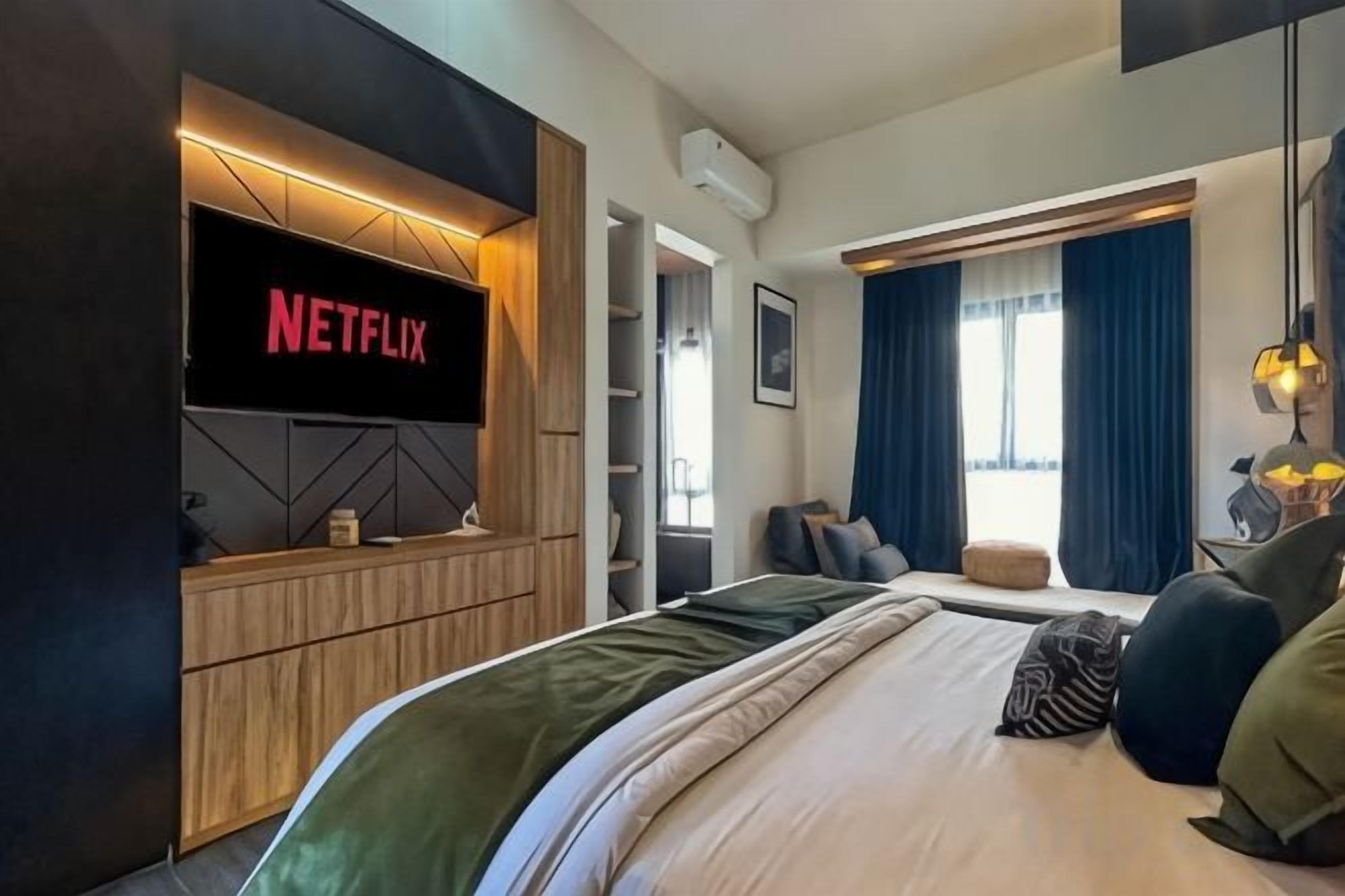 A modern hotel room with a large bed, a flat-screen TV mounted on a wooden panel, a seating area with a sofa, and a window with blue curtains.