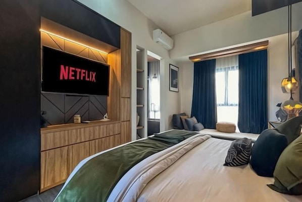 A modern hotel room with a large bed, a flat-screen TV mounted on a wooden panel, a seating area with a sofa, and a window with blue curtains. - Landmark Villas (Bali)