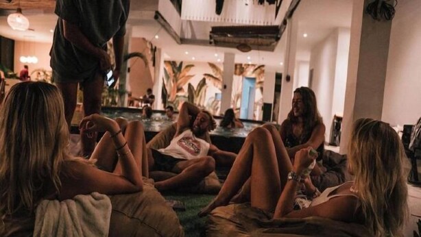 A group of people sitting on bean bags in a spacious, well-lit room with a large window and a view of palm trees. - Coco & Pineapple Pants Hostel (Bali)
