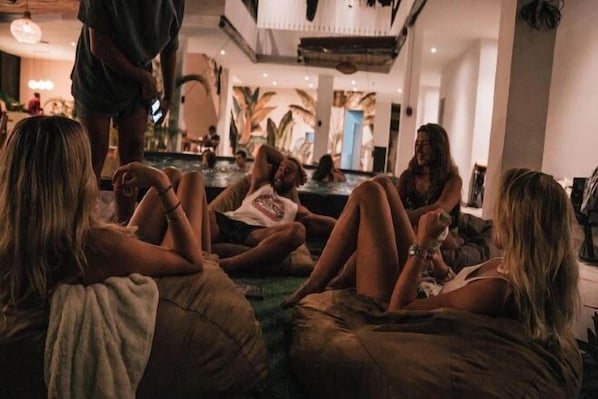 A group of people sitting on bean bags in a spacious, well-lit room with a large window and a view of palm trees. - Coco & Pineapple Pants Hostel (Bali)